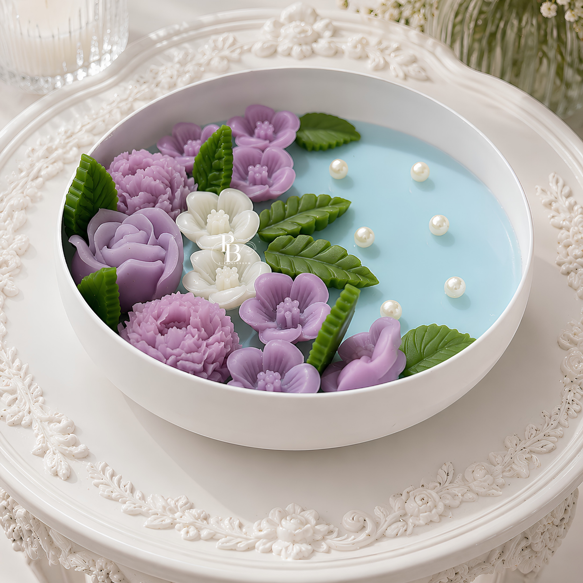 Scented candle in white bowl with blue wax, purple blossoms, green leaves, and pearls
