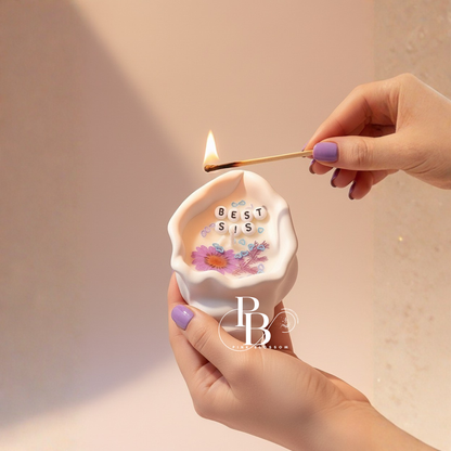 Shell-shaped candle being lit, with floral “BEST SIS” design on the front